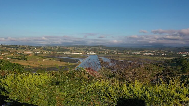 Ripresa di un drone che decolla dove si vede l'ombra del drone e del suo pilota davanti a una recinzione e dietro una foresta e un estuario con vegetazione e case rurali in Cantabria, Spagna.