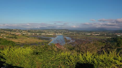 Shot-of-a-drone-taking-off-where-you-can-see-the-shadow-of-the-drone-and-its-pilot-in-front-of-a-fence-and-behind-a-forest-and-estuary-with-vegetation-and-rural-houses-in-Cantabria,-Spain