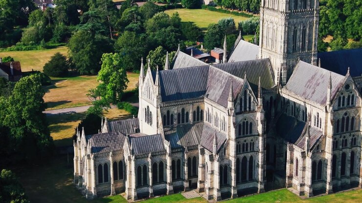 Close drone view of Salisbury Cathedral showing Gothic windows, stone arches, steep roofs, towers and detailed medieval architecture surrounded by green grounds.