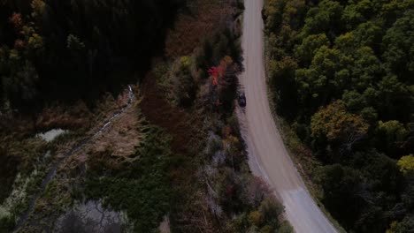 Descending-aerial-of-car-driving-on-dirt-road-through-trees-in-autumn