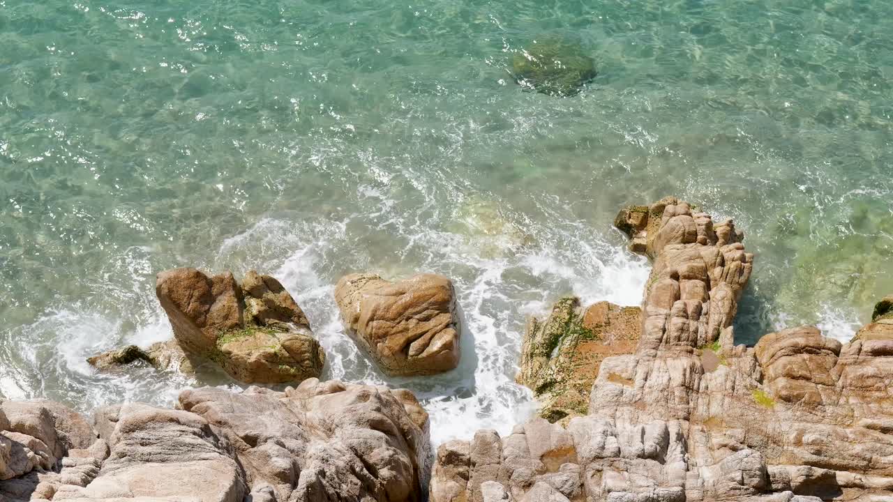 Top-down view of waves crashing against rugged coastal rocks in clear shallow sea water under bright sunlight.