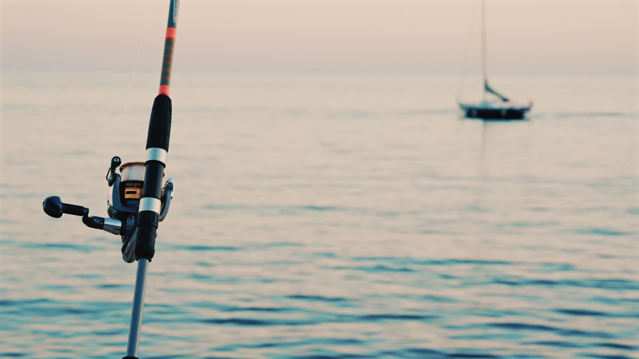 Primo piano di una canna da pesca e un mulinello posizionati vicino al mare, con una barca sfocata sullo sfondo