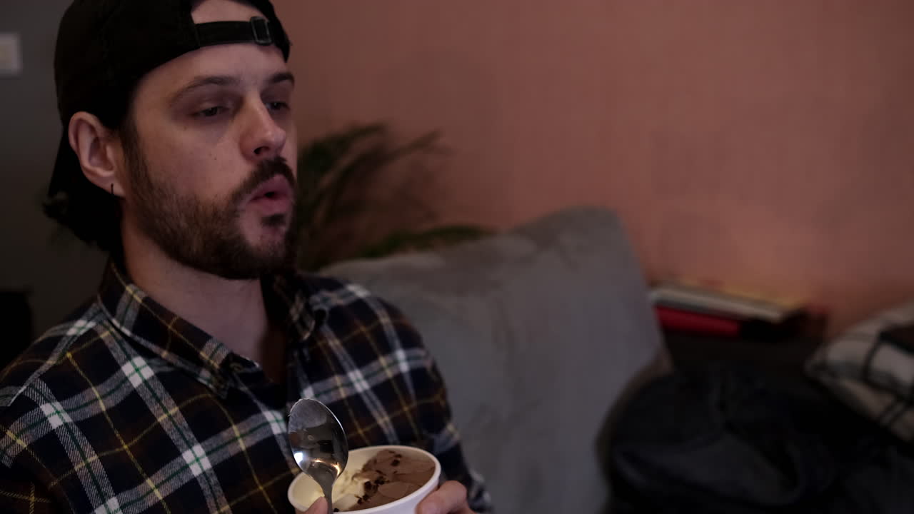 cámara lenta del hombre caucásico que come helado en el sofá, frente a la televisión, en su tiempo libre