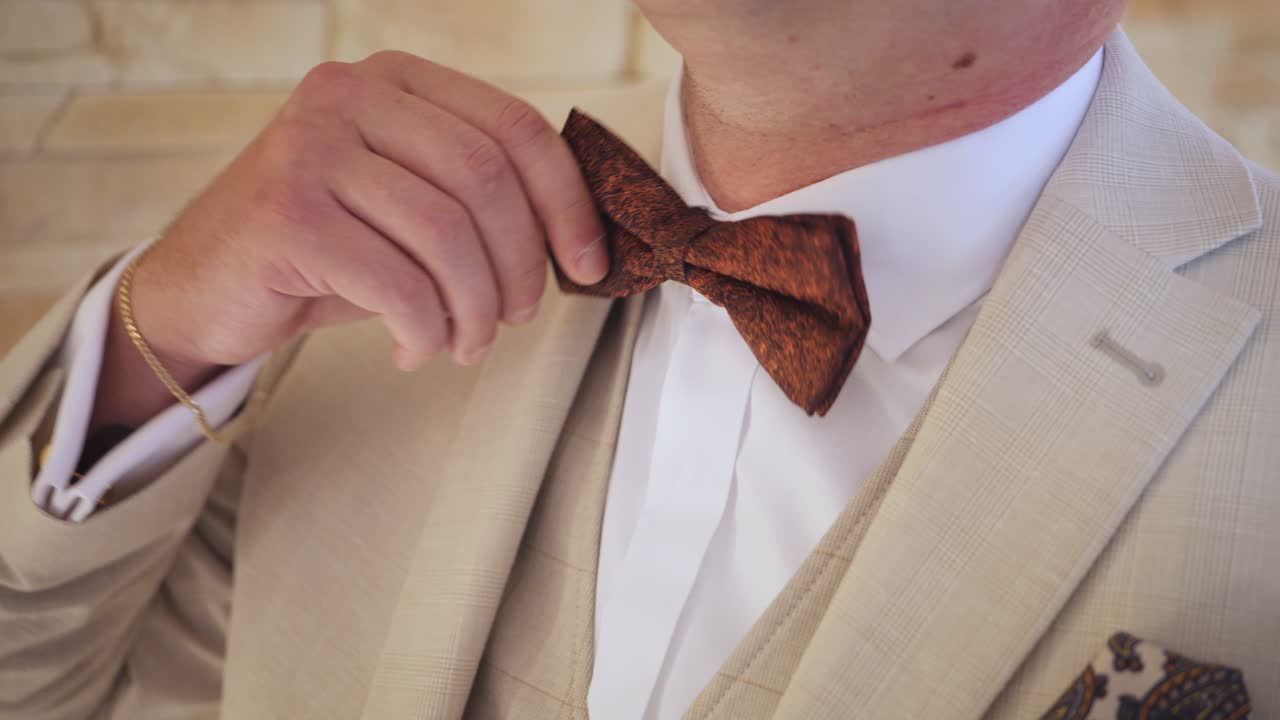 Man adjusts brown bow tie on beige suit for a formal event indoors