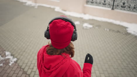 back view of woman in red beanie and hoodie touching headphones while standing on an interlocked path, snow-covered ground and intricate fence design in the background