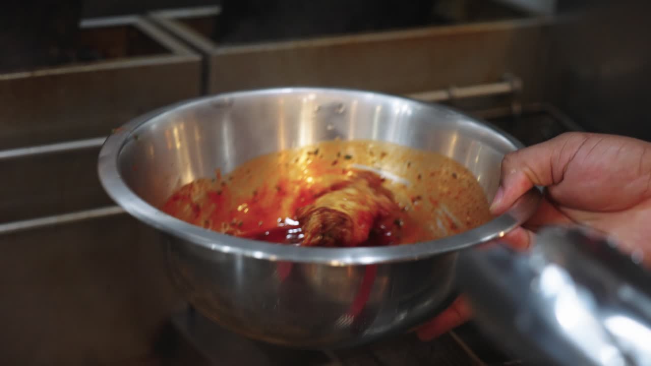 Filmato ravvicinato 4K di ali di pollo mescolate e ricoperte di salsa all'interno di una ciotola per mescolare in acciaio inossidabile in una cucina professionale durante la preparazione del cibo.
