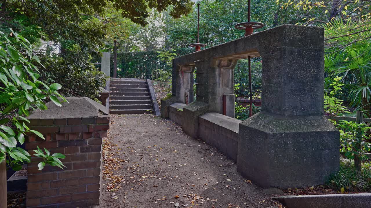 Stone steps lead up behind the historic concrete gate structure of the Jinzaemon Weir in a lush, wooded park setting