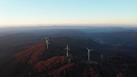 Tranquila-Vista-Aérea-De-Turbinas-Eólicas-En-Lo-Alto-De-Colinas-Cubiertas-De-Bosques-Durante-El-Crepúsculo-Temprano