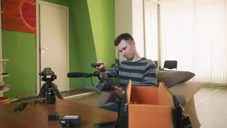 man reaching into camera equipment bag to retrieve microphone in casual workspace. green walls, wooden table with camera gear like lens and tripod visible. cozy setup for video production