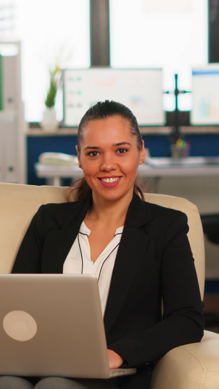 Vertical video Hispanic business woman smiling at camera sitting on couch typing on computer