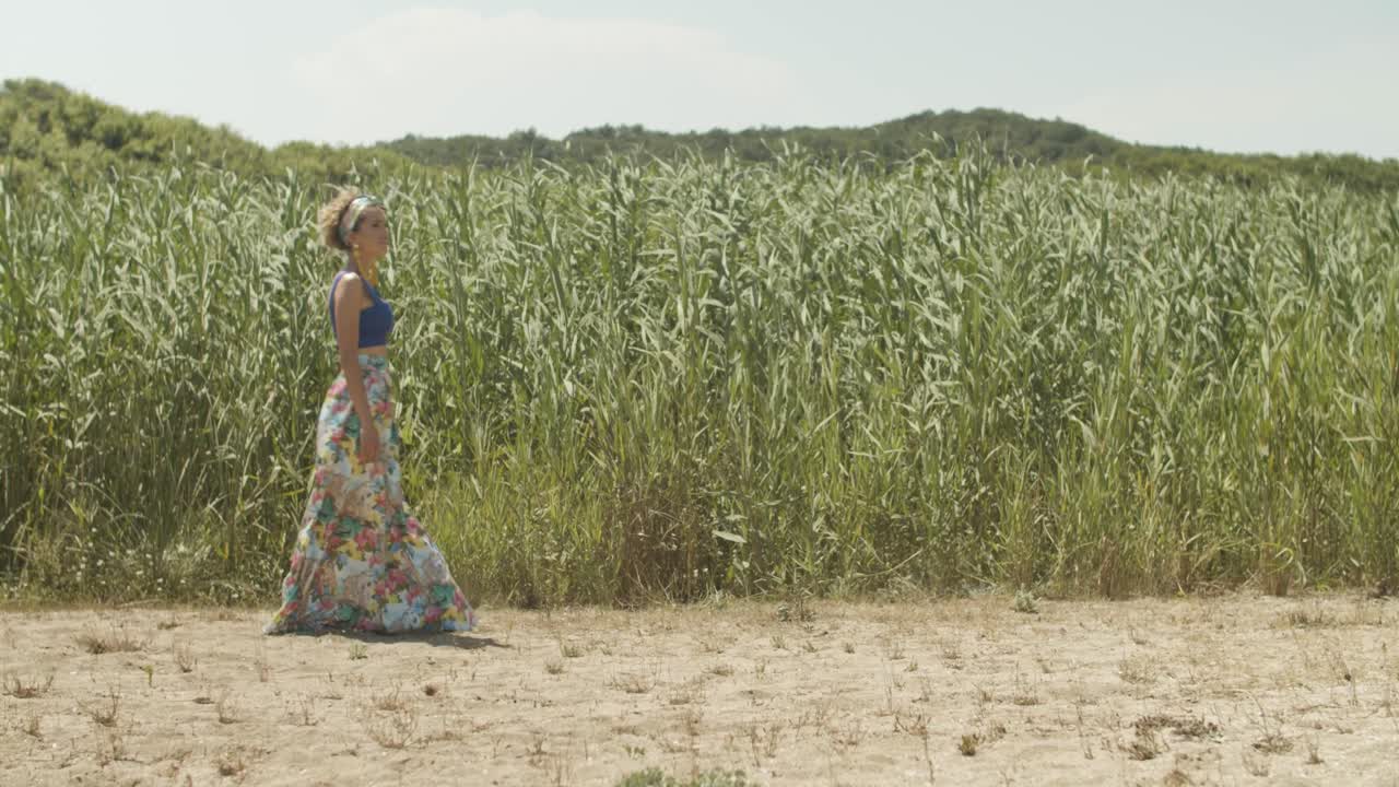 Pretty woman in summer skirt walks leisurely over beach, swaying reed background
