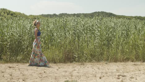 Pretty-woman-in-summer-skirt-walks-leisurely-over-beach,-swaying-reed-background