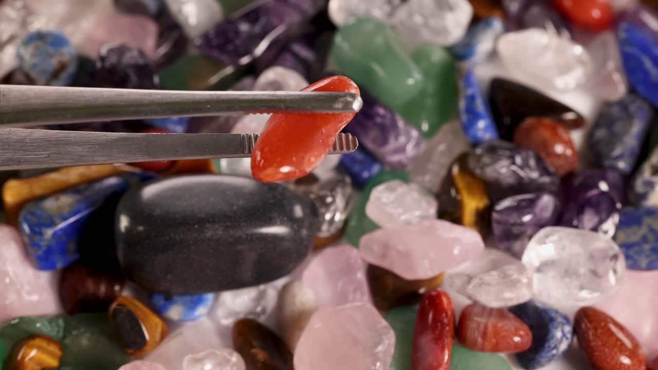 Tweezers carefully pick a red jasper stone from a colorful array of chakra stones under bright lighting