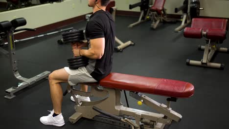 side shot of teen bodybuilder adjusting himself on a bench and lifting a heavy dumbbell over his head