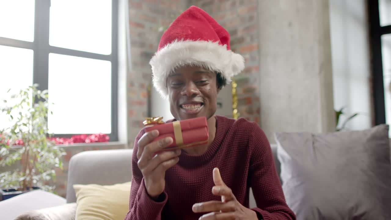 feliz hombre afroamericano con sombrero de santa sentado en el sofá haciendo una llamada de video en casa, en cámara lenta