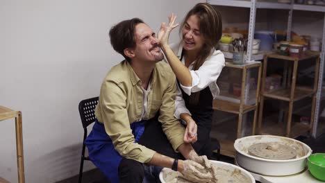 Pareja-Alegre-Y-Sonriente-Divirtiéndose-Mientras-Trabajan-Juntos-En-El-Taller-De-Alfarería.-Sentada-Junto-Al-Torno-De-Alfarero,-Una-Mujer-Tratando-De-Limpiar-La-Cara-De-Su-Novio,-Riendo,-Emocionada.-Estudio-Taller