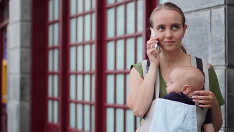 Mientras-Usa-Una-Mochila-Canguro-Para-Llevar-A-Su-Bebé,-Una-Joven-Toma-Fotografías-Con-Un-Teléfono-Móvil-Durante-Sus-Viajes.-Ella-Camina-Y-Revisa-La-Pantalla-Del-Teléfono