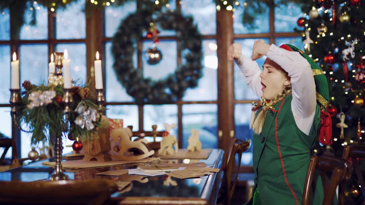 Elf girl woke up at table with letters of congratulations, gifts and candles on the background of the Christmas tree, garlands and a New Year's wreath near the window. Santa Claus assistant stretched and fell asleep again.