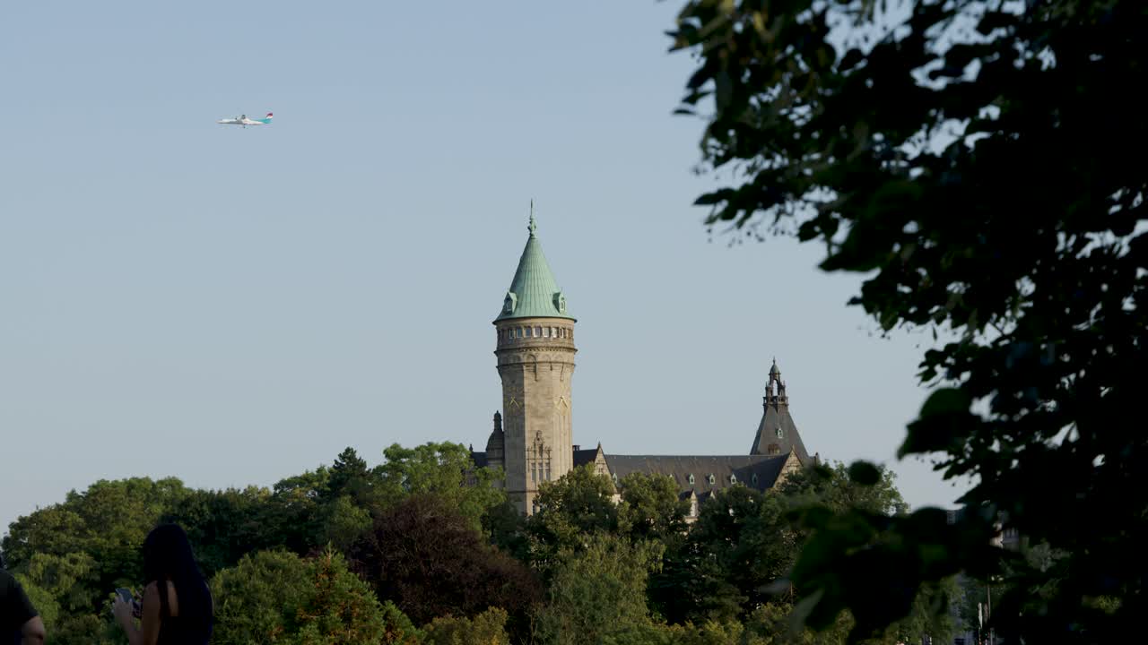 Camera pans past leafy branches to reveal historic tower and spire under clear daylight sky