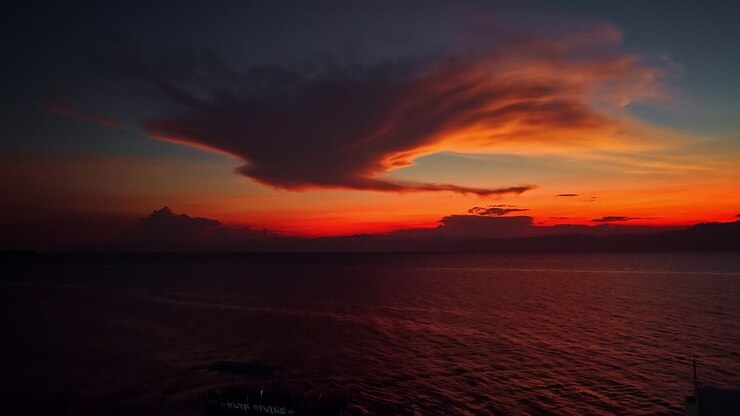 A striking cloud formation hovers over the ocean at sunset, with deep reds and oranges painting the sky