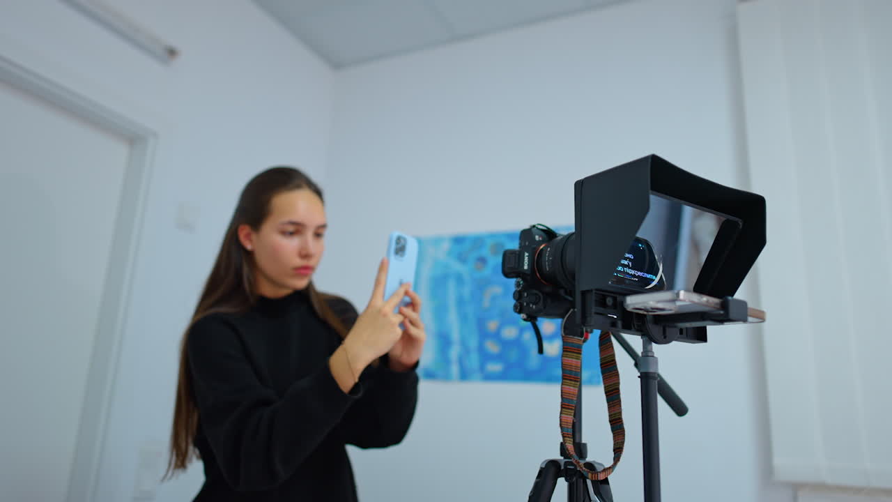 Caucasian long-haired girl wearing black sweater taking video on her smartphone. Lady stands near the professional camera filming in studio.