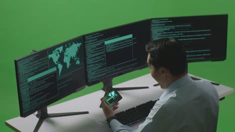 side view of happy asian man developer looking at code on mobile phone while write code with multiple computer screens in green screen studio