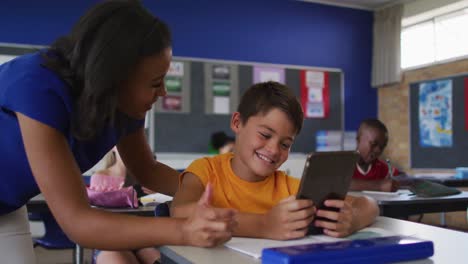 diversa maestra sonriente ayudando a un escolar sentado en el aula, usando una tableta