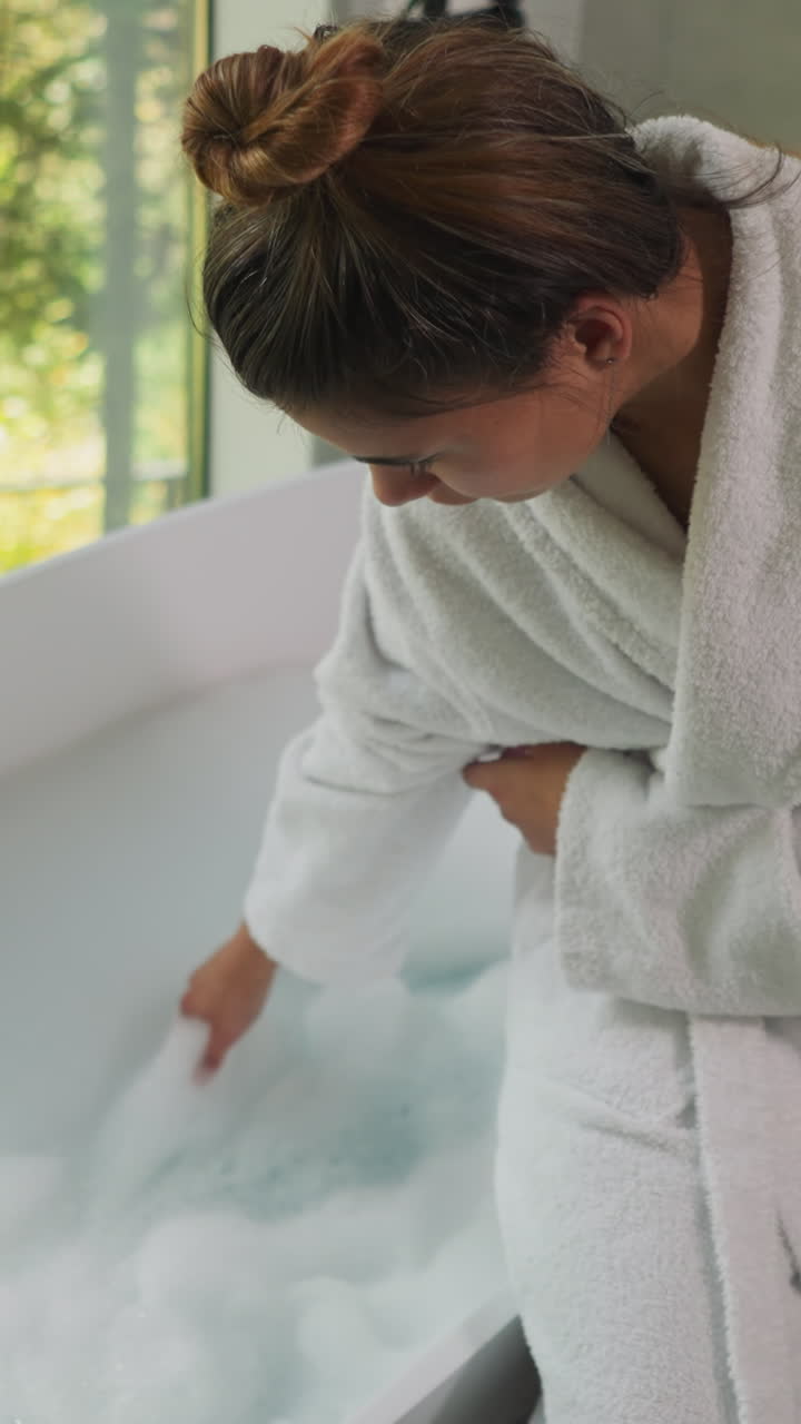 Woman playfully touches froth in bathtub. Lady in terry bathrobe stirs bubble foam checking temperature of water. Preparation for spa procedure