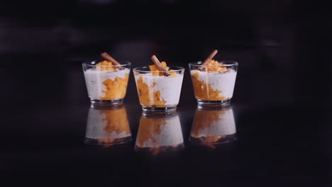 Vasos-De-Panna-Cotta-De-Mango-Dulce-Con-Palitos-De-Canela-Para-El-Postre,-Aislados-En-La-Mesa-De-Cristal-De-Fondo-Negro---Tiro-De-Zoom-Lento