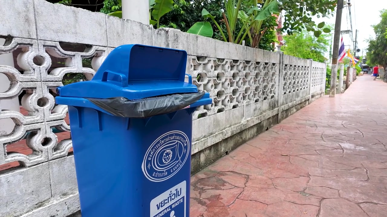 La cámara se acerca a un contenedor de basura azul en un sendero pavimentado junto a una pared blanca.