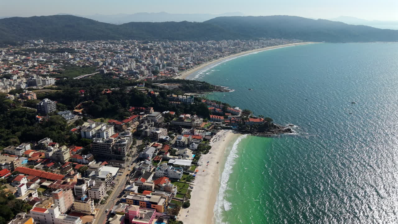 Pristine white sand bordering turquoise Atlantic waters, Bombas Beach nestled between verdant hills and colorful Bombinhas urban landscape