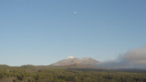 awe inspiring view over mount teide with moon, blue sky and clouds surrounded by green forest