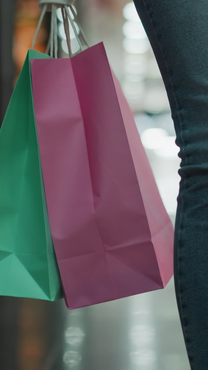 vista de cerca de la pierna de una mujer en vaqueros y zapatillas de lona que lleva bolsas de compras de menta y rosa en su mano derecha mientras camina a través de un centro comercial brillantemente iluminado con pisos de baldosas brillantes