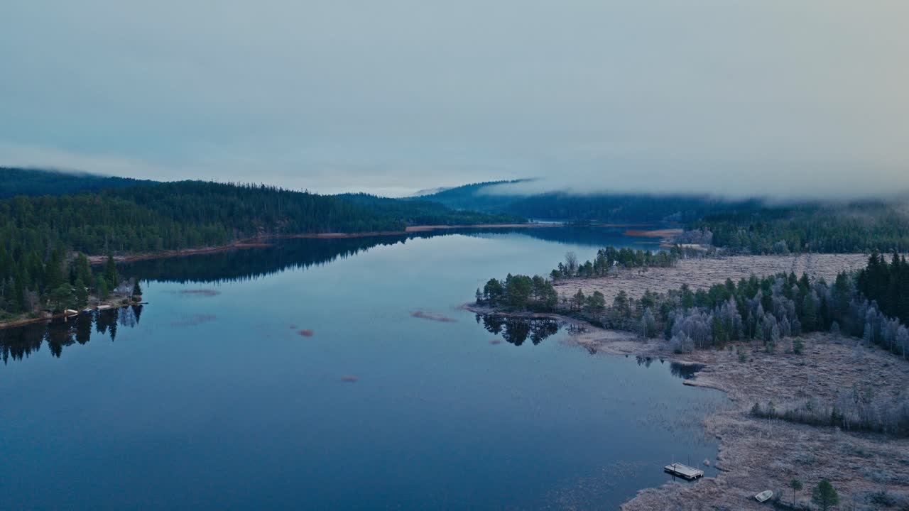 Un lago calmo si estende tra rive boscose sotto un cielo grigio nebbioso - Ripresa aerea con drone