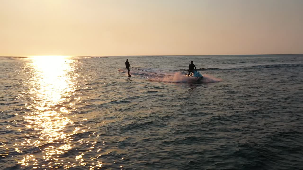 Water sports aerial, silhouette of jet ski pulling man hydro foiling on calm ocean during sunrise