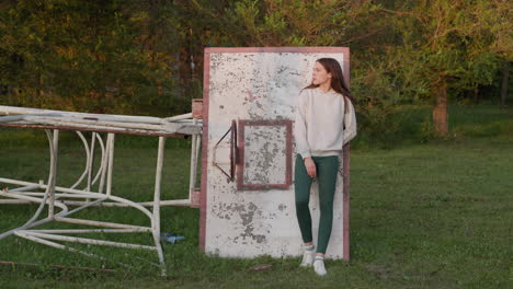 una mujer se apoya en una vieja tabla de baloncesto. una mujer solitaria en ropa deportiva se encuentra entre la hierba verde en una cancha de baloncestro abandonada. un momento de soledad.