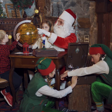santa, little kids and two elves in santa's residence. elves in green costumes playing with old recorder. a boy and a girl looking at globe together with santa.