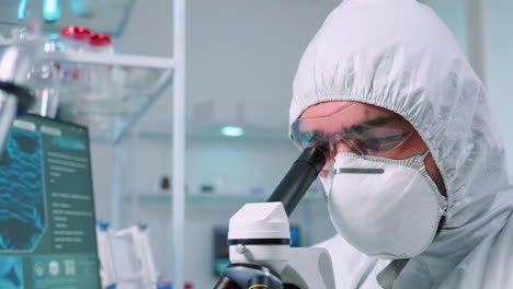 bioquímico en traje de ppe mirando a través de un microscopio durante un experimento