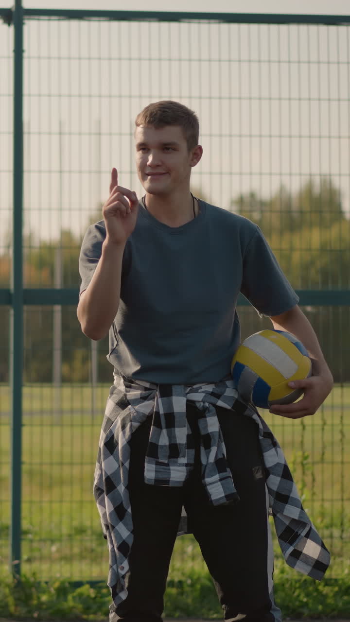 instructor de voleibol con la pelota bajo el brazo apuntando al jugador en el fondo, con la dama con capucha mirando, instalación deportiva en el fondo con entorno al aire libre activo