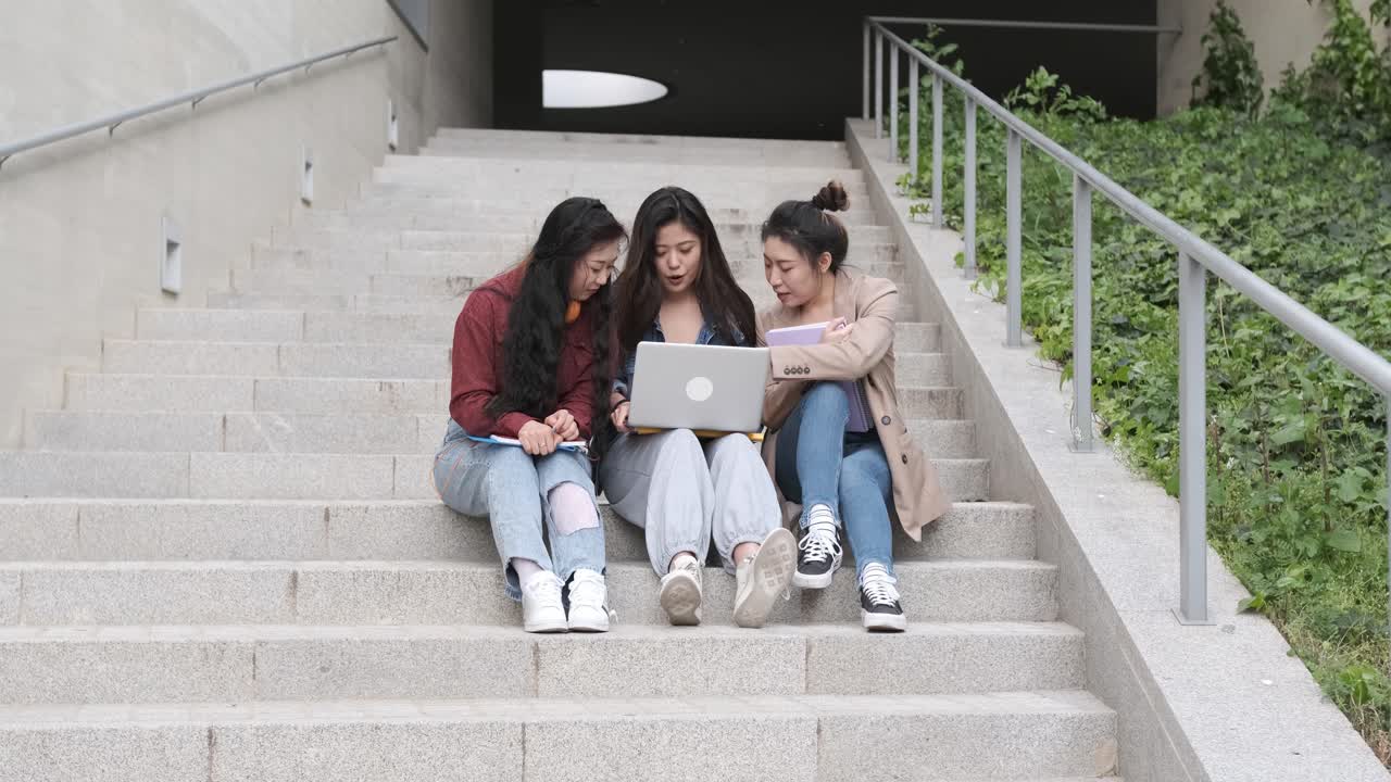 Group of Asian female students using a laptop and studying together while sitting on the stairs of the university campus.