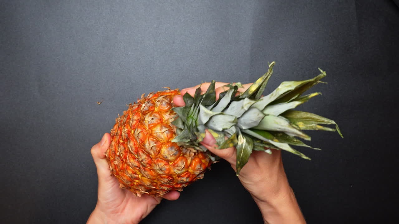 Hands holding a pineapple on a black surface in a kitchen setting. Hands are gripping a pineapple on a dark surface in a kitchen