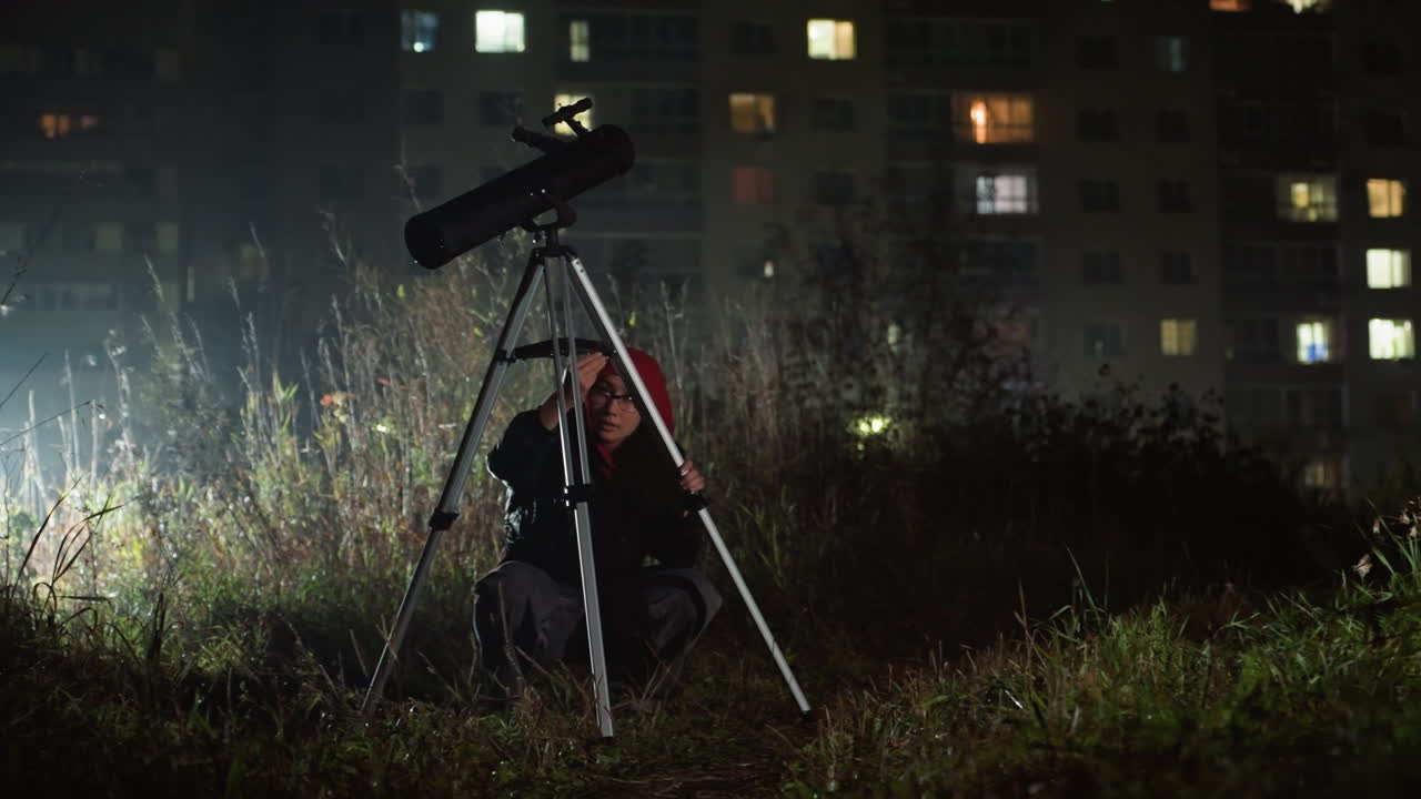 astronomo urbano notturno con telescopio che osserva lo skyline della città e le luci degli appartamenti, treppiede installato nell'erba incolta, bagliore nebbioso, hobbista che regola l'oculare, tranquilla curiosità ed esplorazione paziente