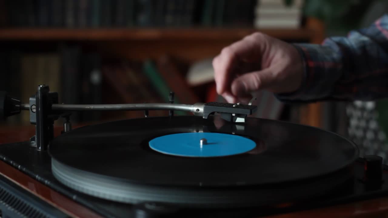 Primo piano di una mano che appoggia delicatamente il braccio di un giradischi su un disco in vinile in rotazione in una stanza accogliente e piena di libri. La scena evoca una connessione nostalgica con la musica analogica, accentuata dalla camicia di flanella casual e
