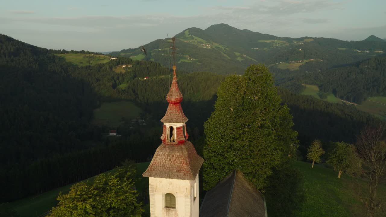 St Thomas Church in Slovenia with birds flying past, surrounded by rolling hills at sunset