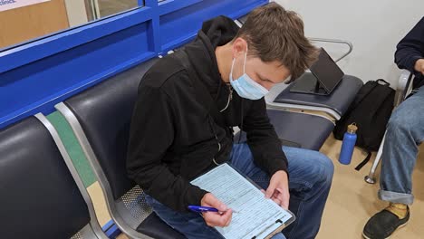 Turista-Con-Mascarilla-Llenando-Un-Papeleo-Médico-Mientras-Espera-En-El-Vestíbulo-De-Un-Hospital