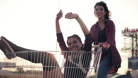 Young-Woman-Is-Sitting-In-The-Grocery-Cart,-While-Her-Friend-Is-Pushing-Her-Behind-At-Parking-By-The-Shopping-Mall-During-Sunset