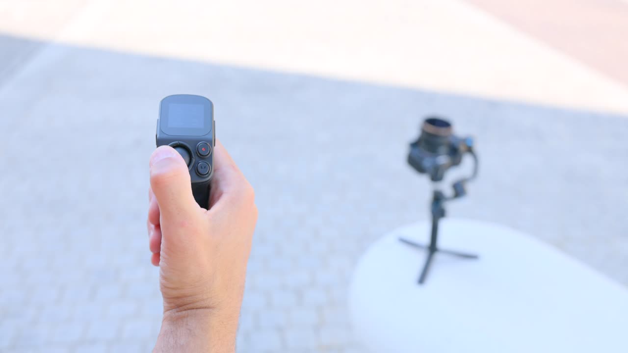 Close-up of hand using remote control handle for FeiyuTech Scorp Mini gimbal. Camera on tripod in blurred background. For technology or filmmaking content