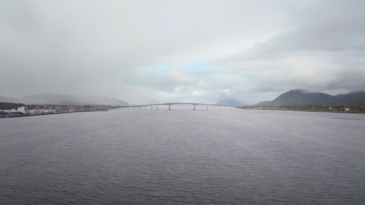 Drone footage over the strait towards Sortland Bridge in Vesterålen. A moody, cloudy day in Northern Norway featuring the town coastline, mountains, and calm sea. Aerial forward, copy space