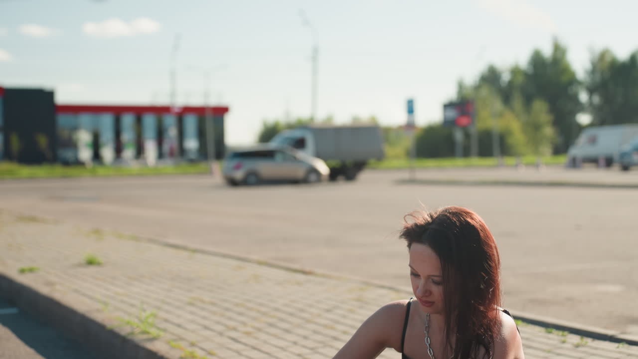 Biker seated on pavement focused on smartphone typing with parked motorcycle in front while cars and vehicles move in busy urban background under bright sunlight