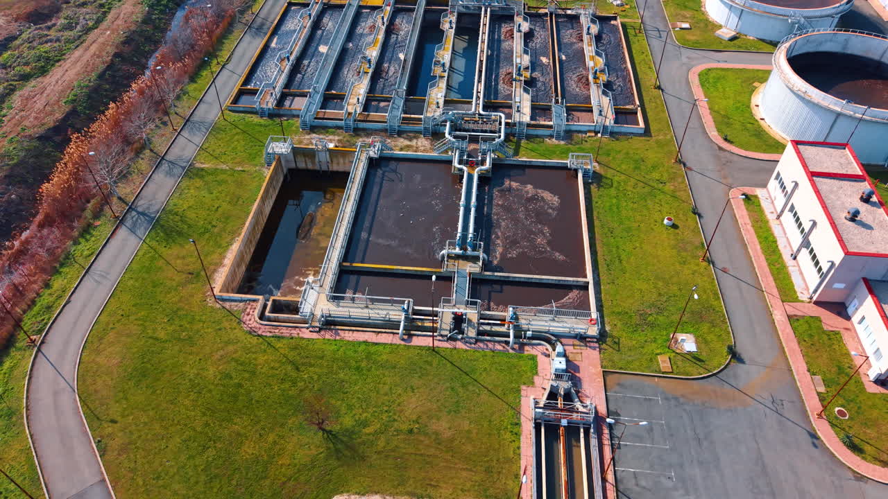 Wide aerial shot of wastewater treatment plant complex. Comprehensive view of sewage facility including tanks, buildings, roads and surrounding green area